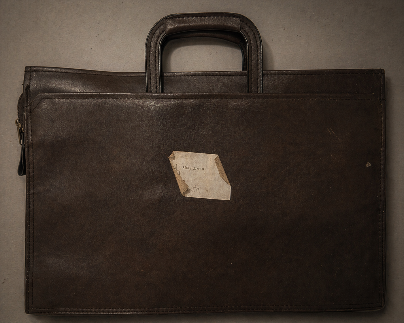 A brown leather briefcase with a hand-labeled manila envelope taped to the front reading 'Red Ricky Simson Hospital' with a drawn red cross